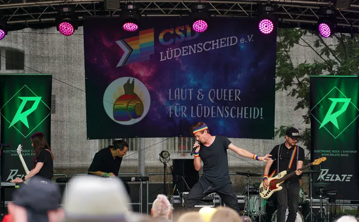 Die Band Relate tritt auf einer Bühne beim Christopher Street Day in Lüdenscheid vor einem Banner mit Regenbogenmotiven auf.