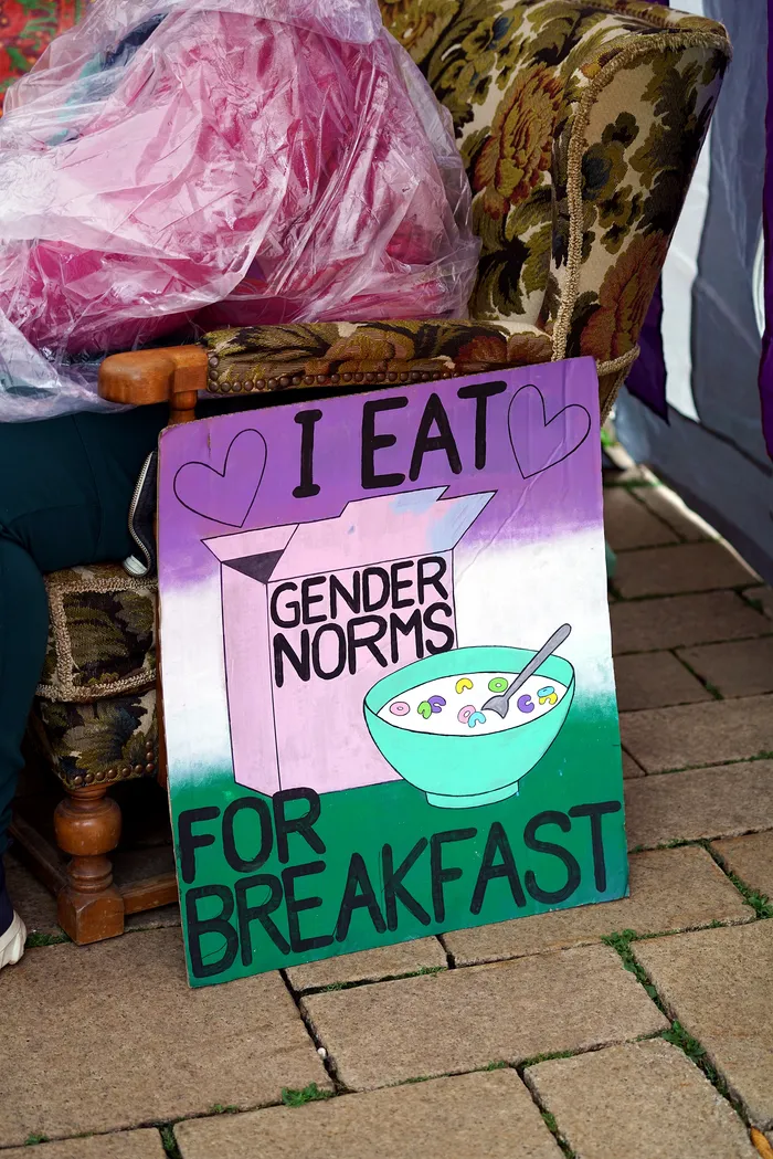 Ein handgemaltes Schild mit der Aufschrift „I EAT GENDER NORMS FOR BREAKFAST“ und Illustrationen eines Kartons und einer Müslischale lehnt an einem gemusterten Sessel.