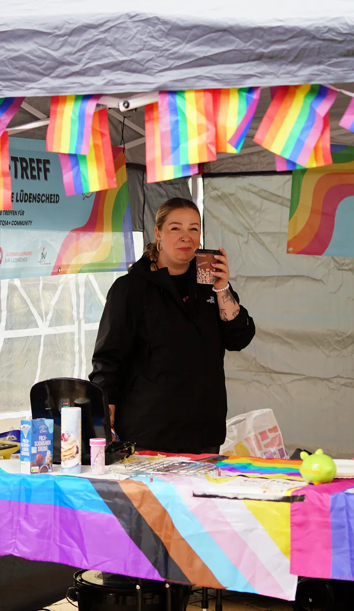 Eine Frau steht hinter einem mit Regenbogenfarben dekorierten Tisch an einem Stand beim Christopher Street Day in Lüdenscheid.