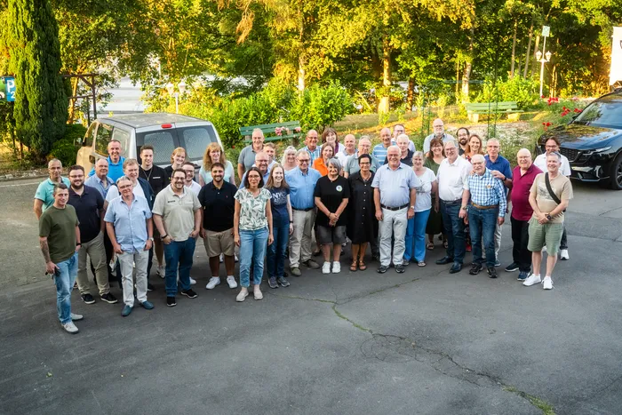 Eine große Gruppe von Menschen posiert für ein Gruppenfoto im Freien vor einer Kulisse aus Bäumen.