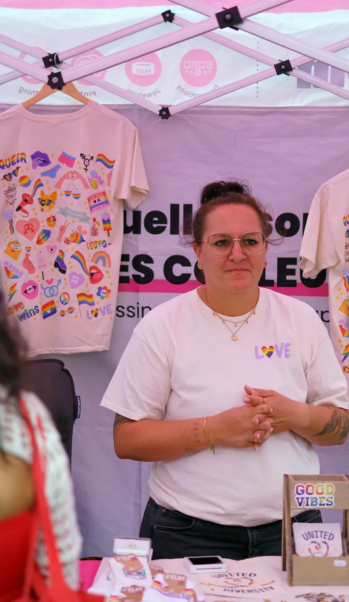 Eine Frau mit Brille steht hinter einem Verkaufsstand mit LGBTQ+-Merchandise; im Hintergrund hängen T-Shirts mit bunten Regenbogen-Motiven.
