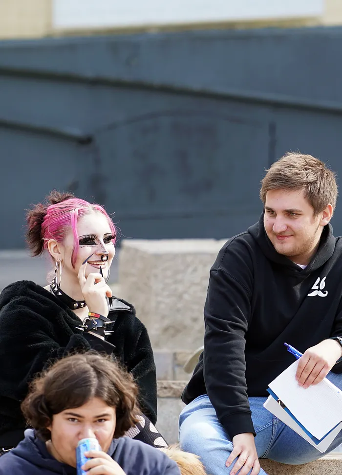 Eine Person mit pinkfarbenen Haaren und dunklem Make-up lächelt einen jungen Mann in einem schwarzen Kapuzenpullover an, der ein Notizbuch hält.