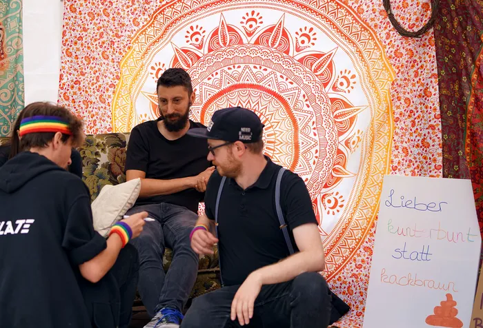 Drei Männer sitzen vor einem großen, orangefarbenen Mandala-Hintergrund. Ein Schild daneben trägt die Aufschrift „Lieber kunterbunt statt kackbraun“.