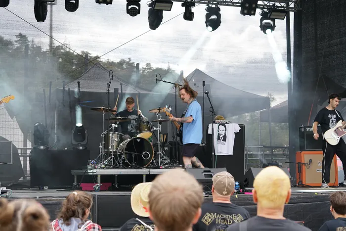 Eine Band tritt auf einer Außenbühne mit Schlagzeug, Bass und Gitarre unter Scheinwerfern und Bühnennebel auf.