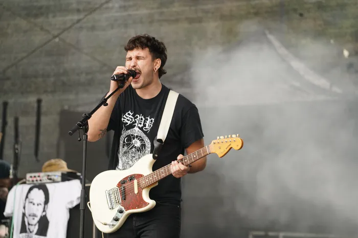 Ein Musiker singt mit geschlossenen Augen in ein Mikrofon und spielt eine weiße E-Gitarre vor einem rauchigen Hintergrund.