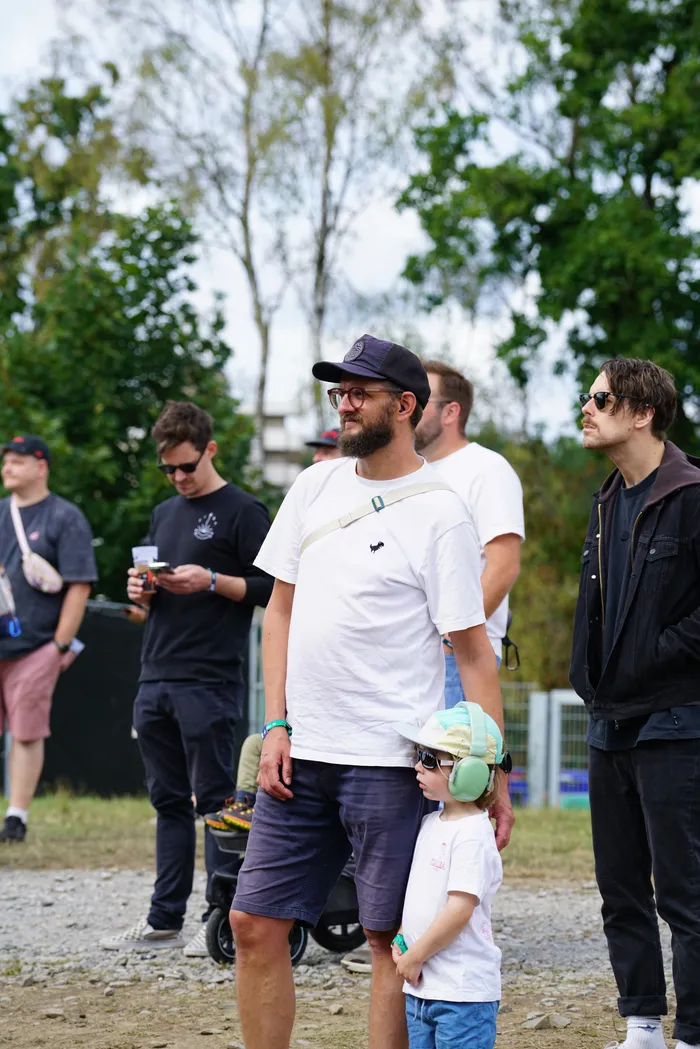 Ein Mann mit Bart und dunkler Kappe in einem weißen T-Shirt steht neben einem kleinen Kind, das Kopfhörer und eine helle Kappe trägt.