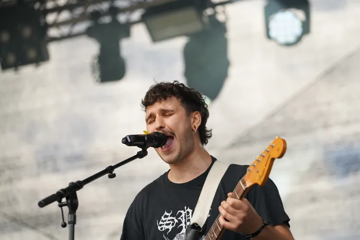 Ein Mann mit dunklen Locken singt mit geschlossenen Augen in ein Mikrofon und spielt eine E-Gitarre.