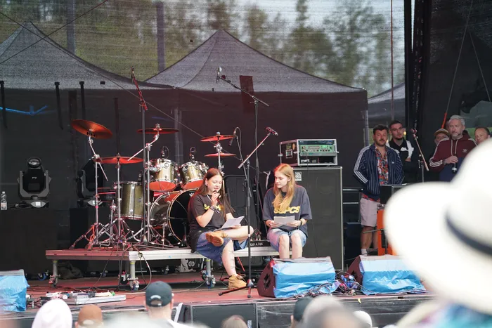 Eine Musikerin spielt Gitarre und singt auf einer Bühne, während eine weitere Frau daneben sitzt und ein Blatt Papier liest. Im Hintergrund stehen ein Schlagzeug und weitere Personen.