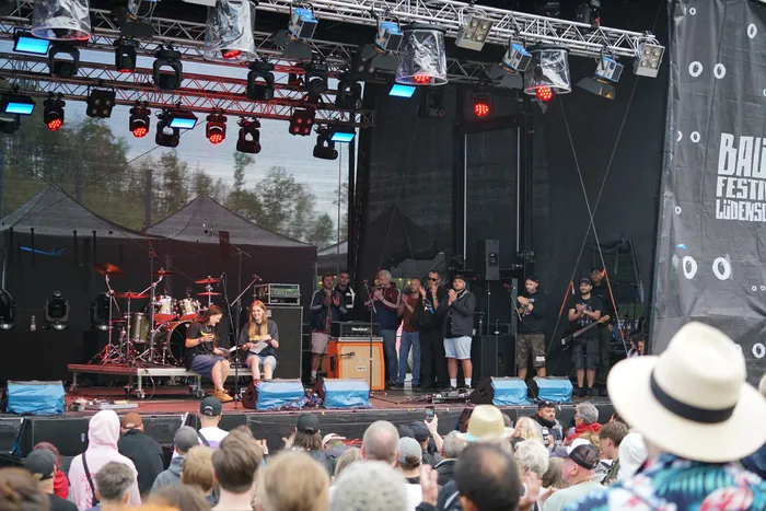 Musiker treten auf einer beleuchteten Außenbühne bei einem Tribut-Konzert vor einem Publikum auf.