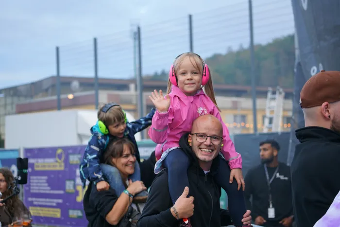Ein lächelnder Mann trägt ein Mädchen mit rosa Kopfhörern auf den Schultern, das in die Kamera winkt.