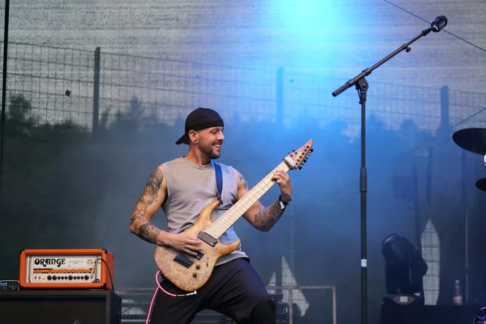 Ein tätowierter Mann mit schwarzer Kappe spielt eine helle E-Bass-Gitarre auf einer blau beleuchteten Bühne.