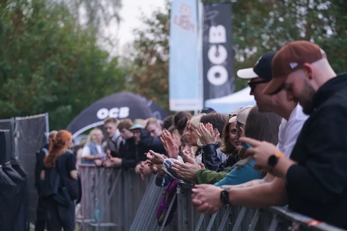 Menschen stehen an einer Metallabsperrung und klatschen bei einem Event. Im Hintergrund sind Banner und ein Zelt mit der Aufschrift „OCB“ zu sehen.