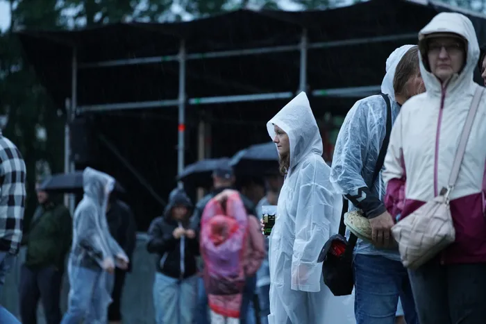 Menschen in hellen Regenponchos stehen bei Regen vor einer dunklen Bühnenkonstruktion.