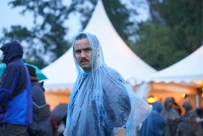 Ein Mann mit Schnurrbart in einem blauen, transparenten Regenponcho vor weißen Festzelten.