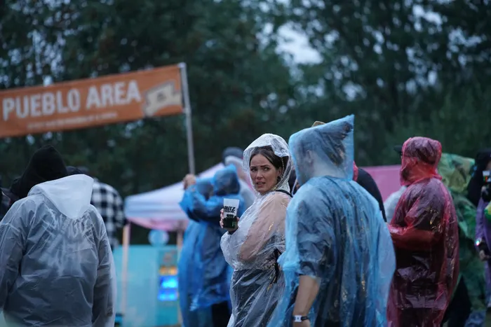 Eine Frau in einem durchsichtigen Regenponcho hält einen Becher und blickt besorgt. Im Hintergrund stehen Menschen in bunten Regenponchos vor einem „PUEBLO AREA“-Banner.