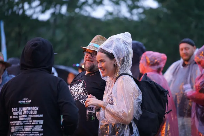 Ein Mann mit Brille und Strohhut sowie eine Frau in einem durchsichtigen Regenmantel stehen bei einem Outdoor-Event.