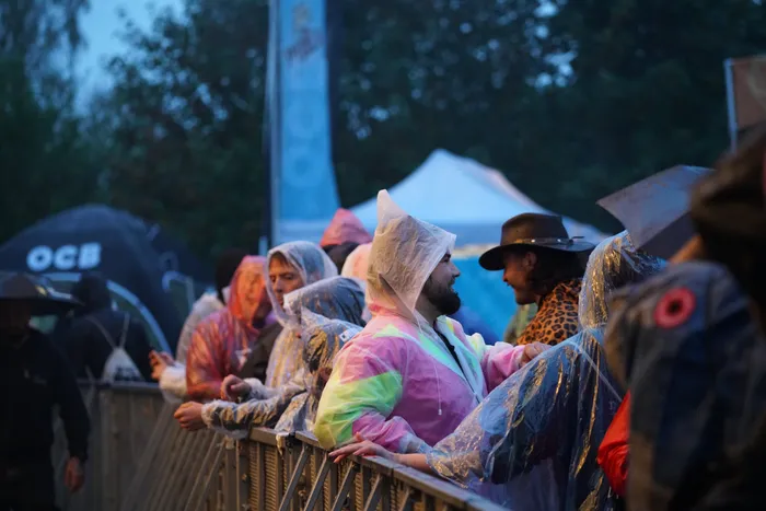 Menschen in bunten und transparenten Regenponchos stehen hinter einer Metallabsperrung bei einem Open-Air-Event.
