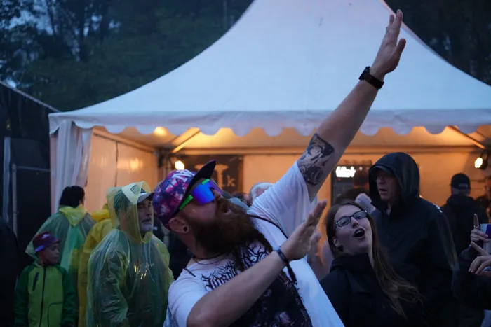 Ein bärtiger Mann mit bunter Kappe und Sonnenbrille hebt die Arme bei einem Event unter einem weißen Zelt.
