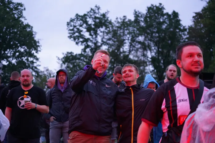 Eine Gruppe von Menschen bei der Bautz26 Collabo Stage, wobei ein lächelnder Mann in einer schwarzen Jacke einen kleinen Gegenstand hochhält.