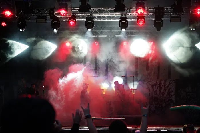 Musiker auf einer Bühne unter roten und weißen Scheinwerfern, umhüllt von dichtem Nebel. Im Vordergrund sind die Silhouetten des Publikums zu sehen.