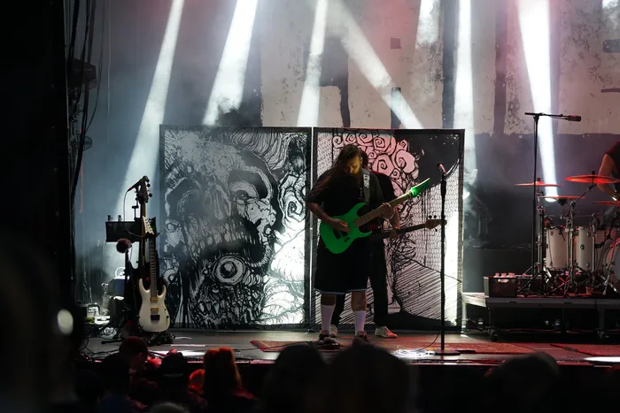Ein Musiker spielt eine grüne E-Gitarre auf einer Bühne vor großformatigen, schwarz-weißen Grafiken und hellen Lichtstrahlen.