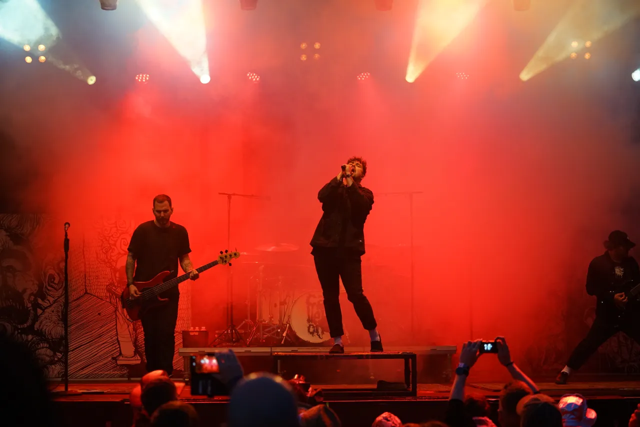 Die Band Callejon performt auf einer Bühne unter intensivem rotem Licht und Nebel. Im Zentrum singt der Sänger, links spielt ein Bassist.