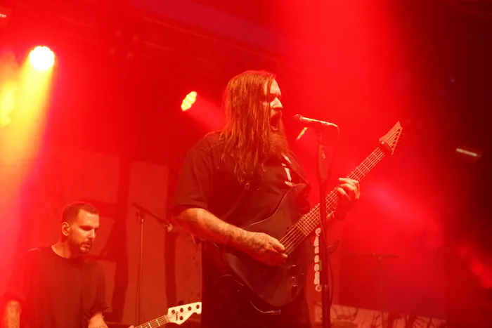 Ein langhaariger Musiker singt in ein Mikrofon und spielt eine schwarze E-Gitarre auf einer intensiv rot beleuchteten Bühne.