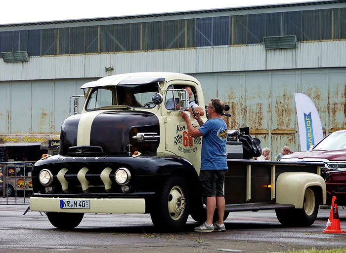 Ein Mann in einem blauen T-Shirt inspiziert einen alten, zweifarbigen Ford-Lastwagen mit einem auffälligen Kühlergrill und großen Scheinwerfern. Im Hintergrund sind weitere Fahrzeuge und ein großes Gebäude zu sehen.