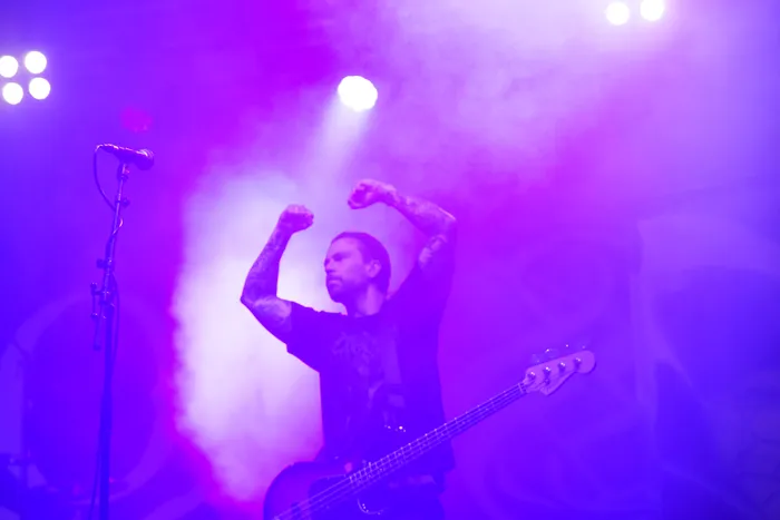 Ein Musiker der Band Sylosis spielt Bass und hebt die Arme unter lila Bühnenlicht und Nebel.