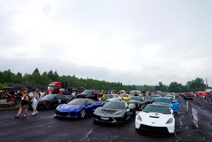 Mehrere Sportwagen, darunter auffällige Corvettes, stehen auf einem asphaltierten Platz vor einer Zuschauermenge und Bäumen.