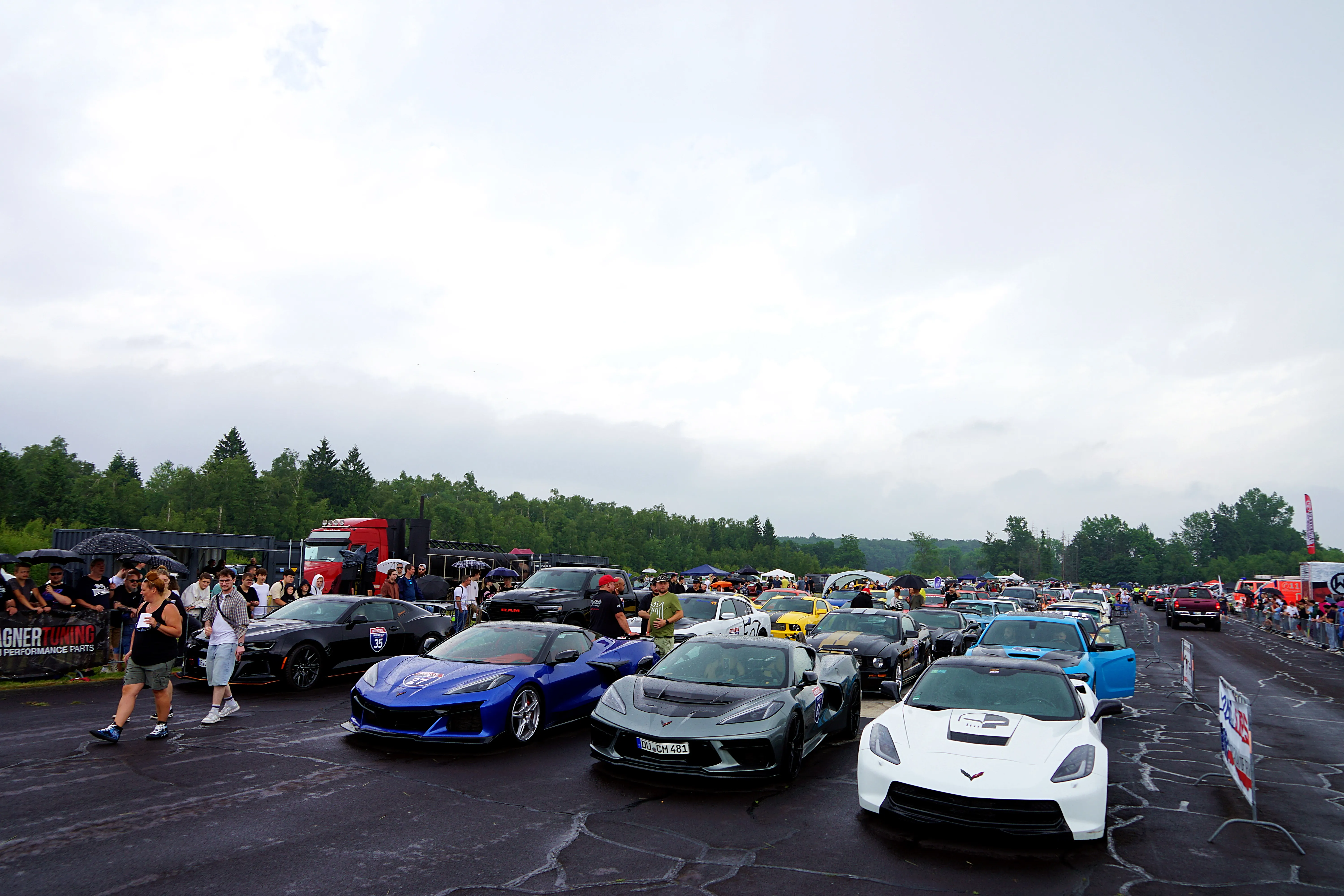 Mehrere Sportwagen, darunter auffällige Corvettes, stehen auf einem asphaltierten Platz vor einer Zuschauermenge und Bäumen.