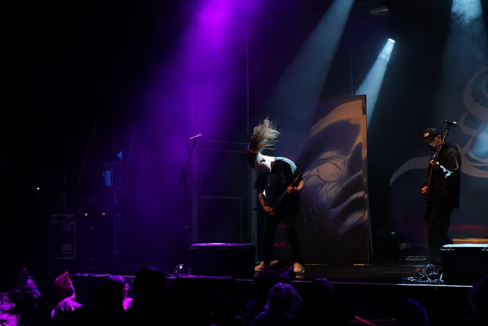 Ein Musiker der Band Sylosis headbangt unter violettem und weißem Bühnenlicht während eines Auftritts.
