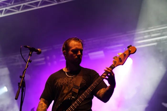 Ein Musiker der Band Sylosis spielt unter lila Bühnenlicht Bassgitarre auf einer Bühne.