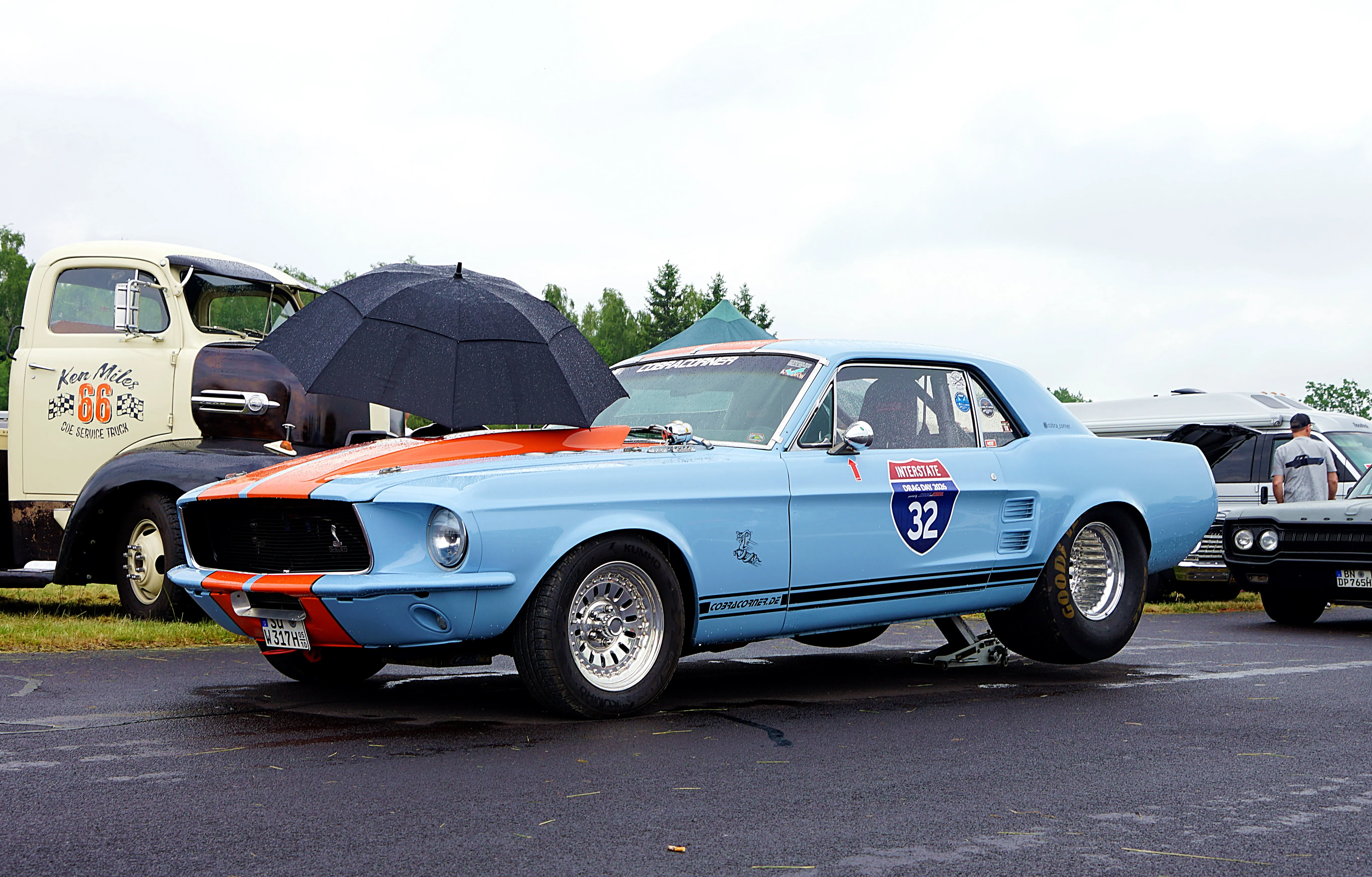 Hellblauer Ford Mustang Fastback mit Rennstreifen und der Nummer 32 auf der Motorhaube steht auf einem Parkplatz, geschützt durch einen geöffneten Regenschirm. Im Hintergrund ein Oldtimer-Lastwagen und weitere Fahrzeuge.
