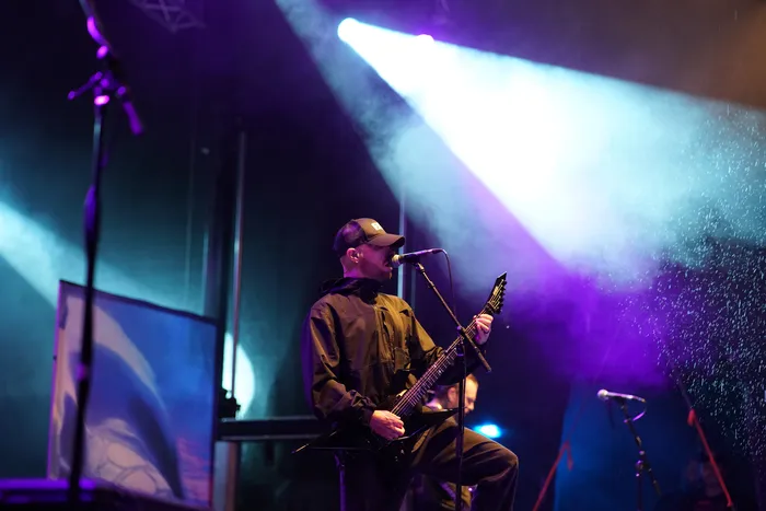 Ein Musiker der Band Sylosis spielt eine schwarze E-Gitarre und singt in ein Mikrofon unter blauem und violettem Bühnenlicht.