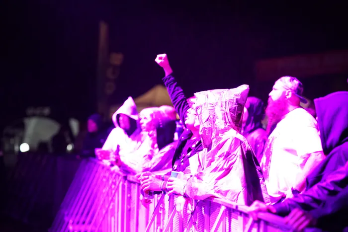 Eine Person mit Kapuze reckt die Faust in die Höhe vor einer Absperrung bei einem Konzert unter lila Beleuchtung.