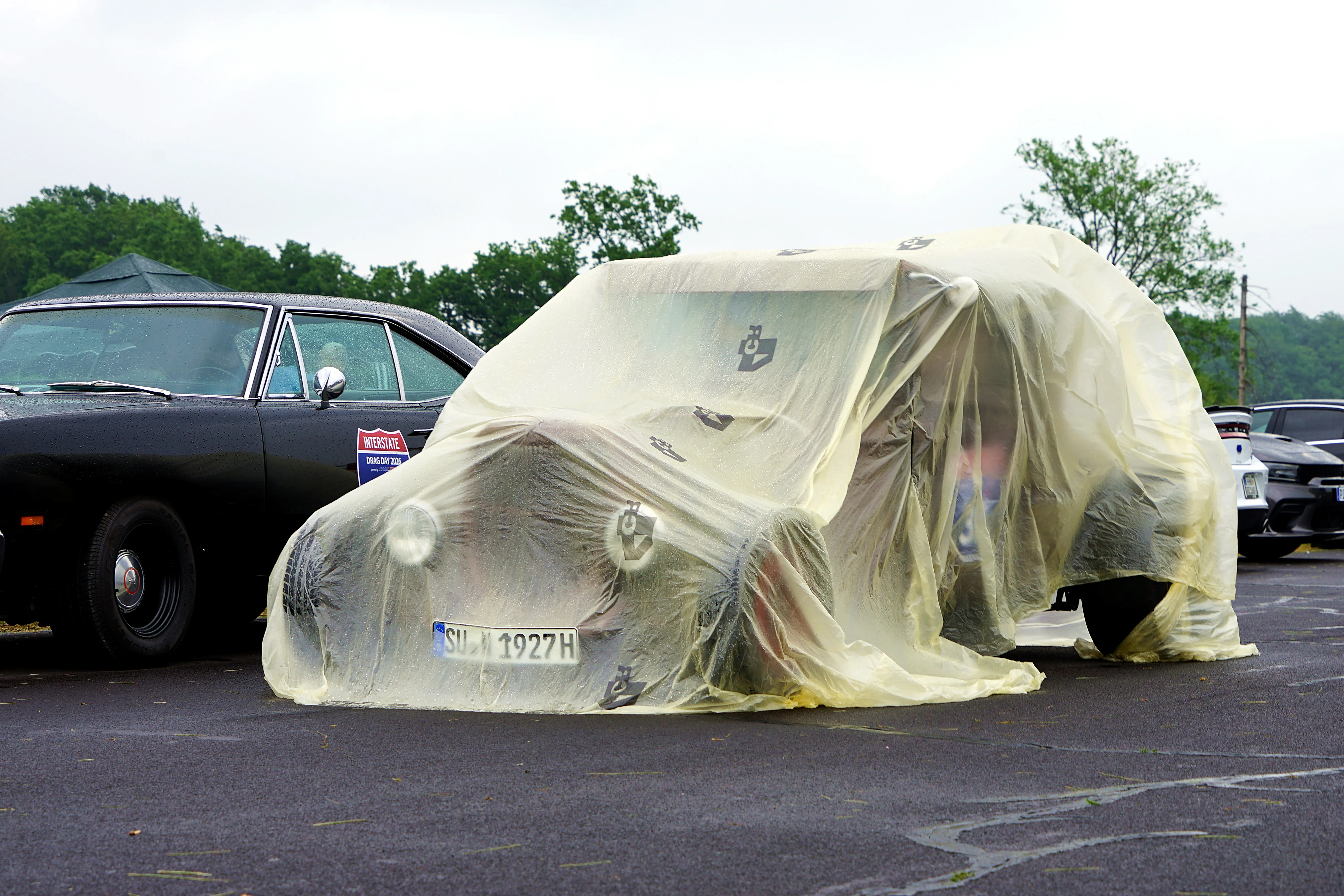 Ein Oldtimer, bedeckt mit einer transparenten Schutzhülle, steht auf einem Parkplatz neben einem dunklen Fahrzeug. Das Kennzeichen des Oldtimers ist teilweise sichtbar.