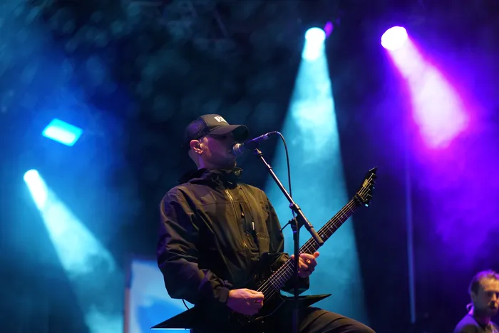 Ein Musiker der Band Sylosis singt in ein Mikrofon und spielt eine schwarze E-Gitarre unter blauem und violettem Bühnenlicht.