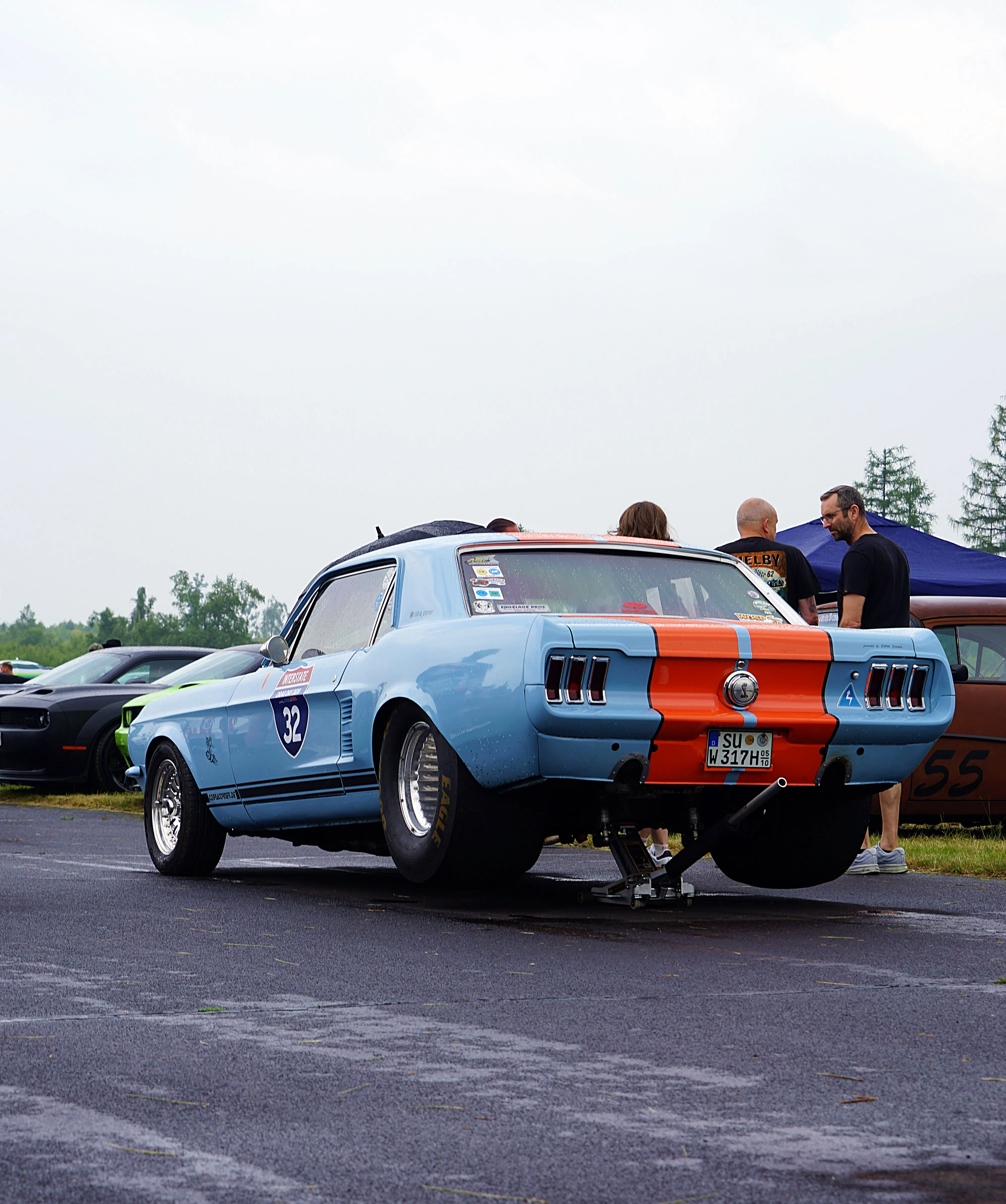 Heckansicht eines hellblauen Ford Mustangs mit Rennstreifen und Startnummer 32, der auf einem Parkplatz steht. Im Hintergrund sind weitere Autos und Personen zu sehen.