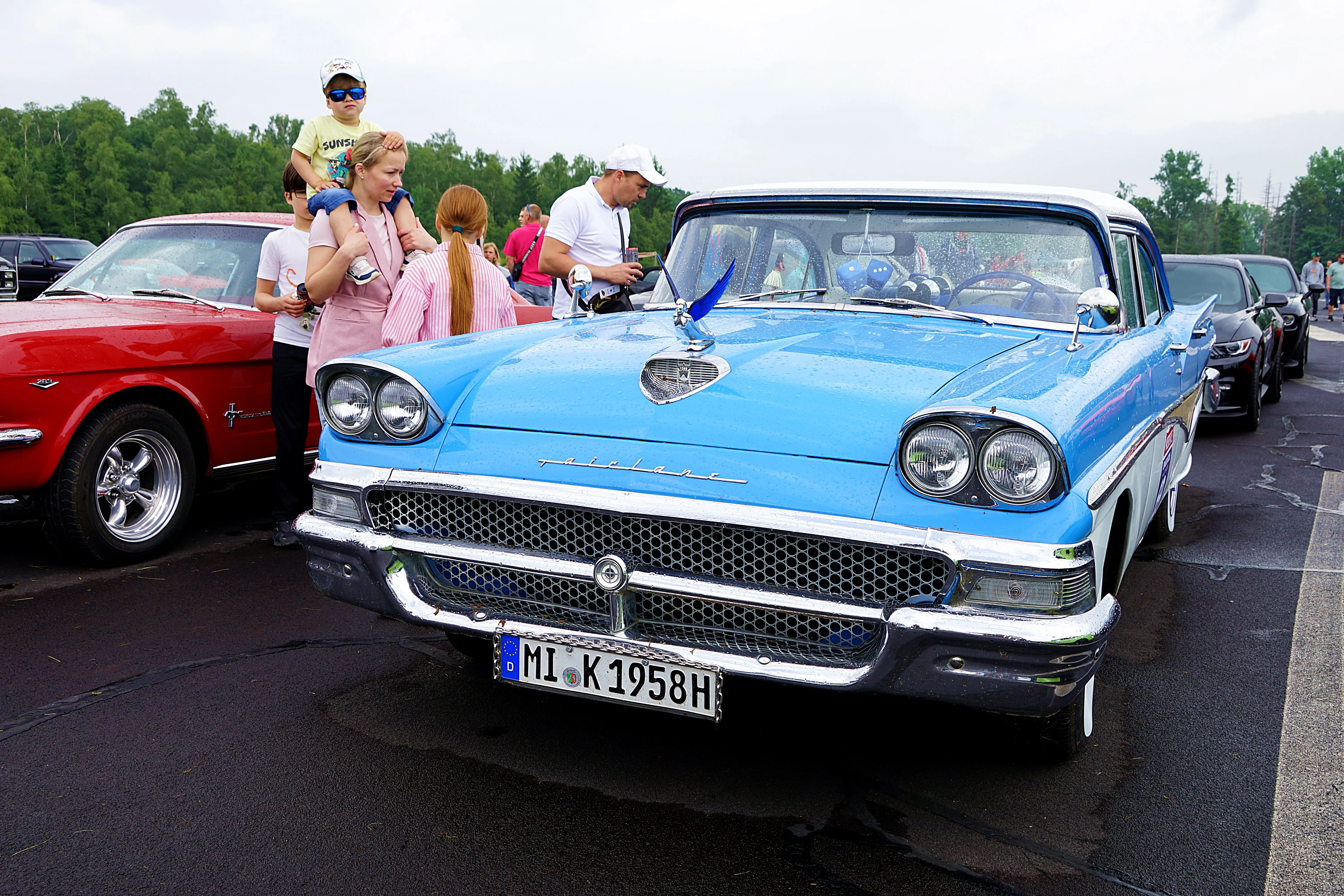 Heller blauer Oldtimer mit Chromdetails steht auf einem Parkplatz, umgeben von weiteren Fahrzeugen und Zuschauern.