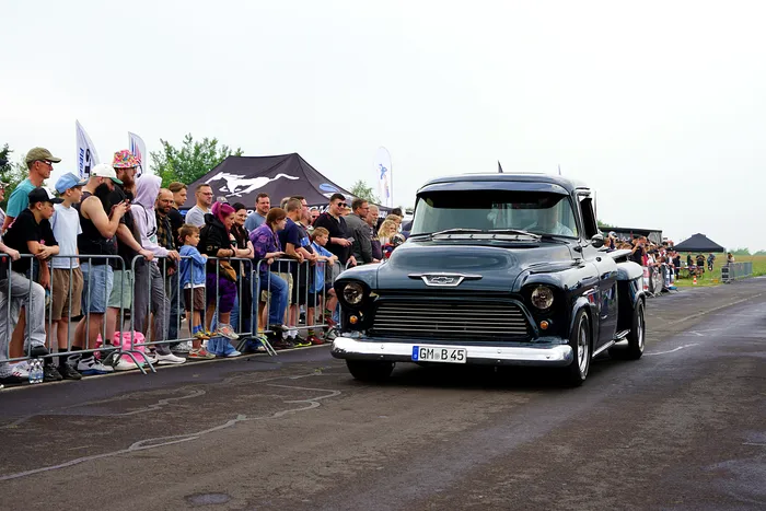 Ein dunkler Oldtimer-Truck fährt an einer Absperrung mit Zuschauern vorbei, im Hintergrund Zelte und weitere Menschen.