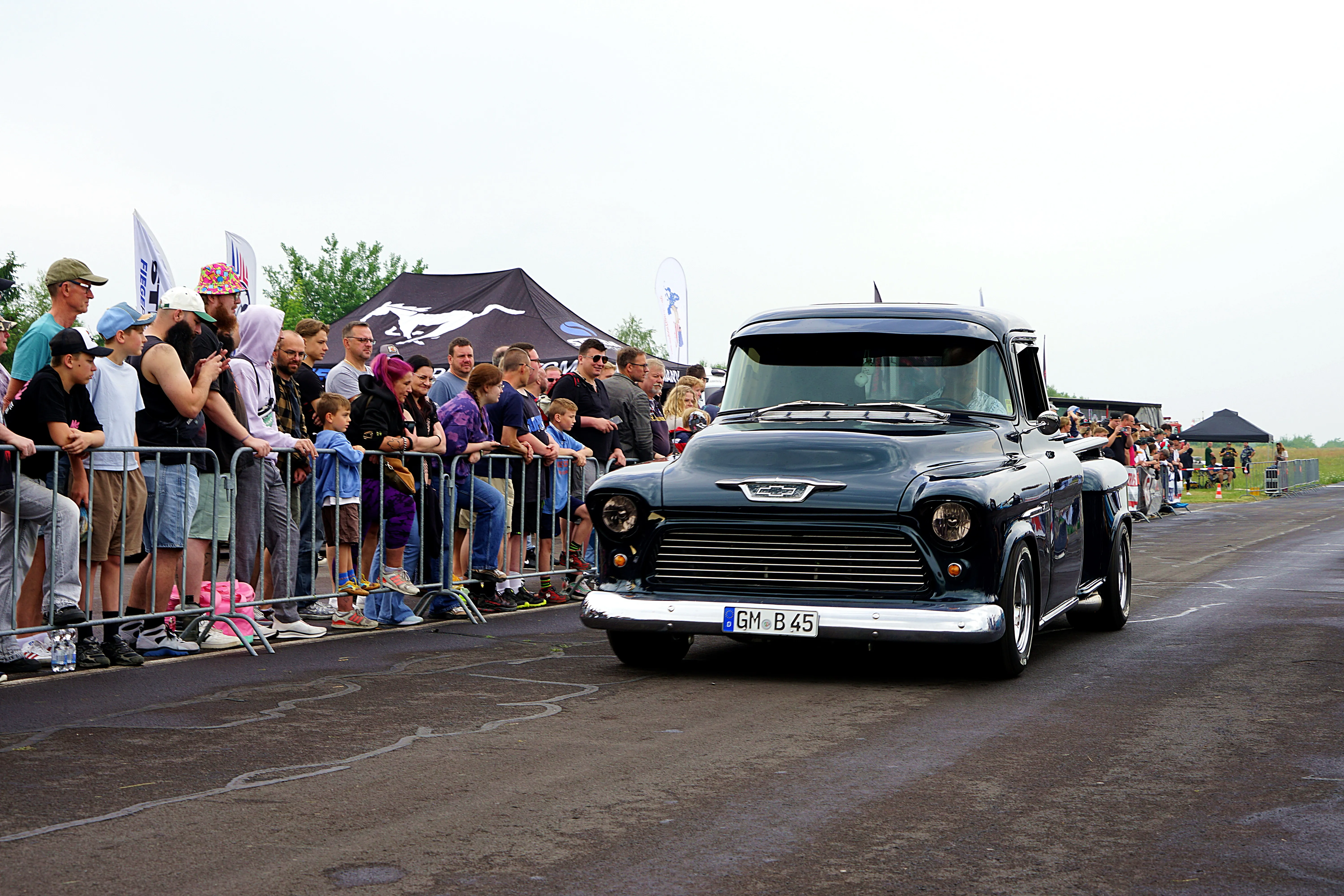 Ein dunkler Oldtimer-Truck fährt an einer Absperrung mit Zuschauern vorbei, im Hintergrund Zelte und weitere Menschen.