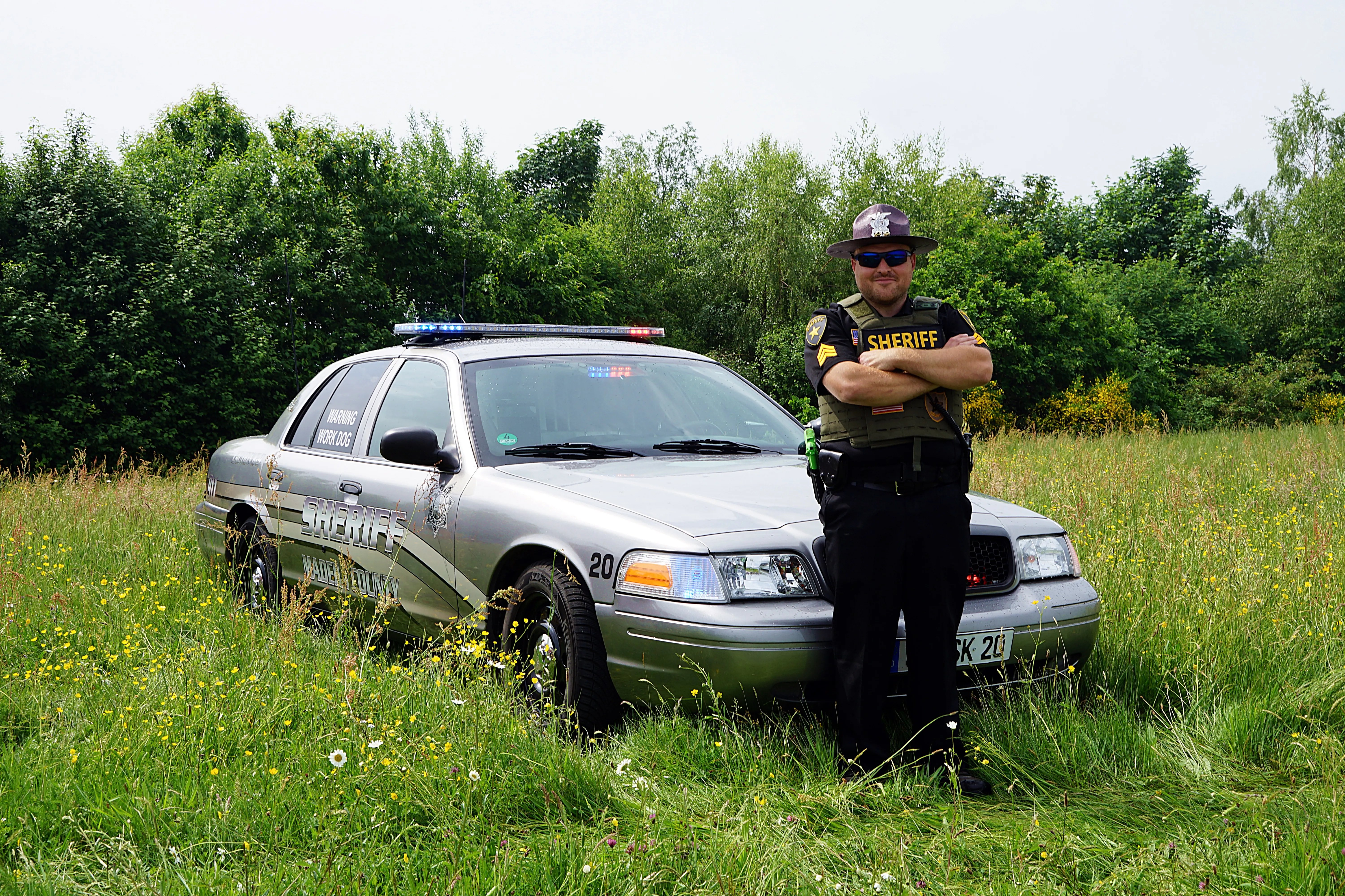 Ein Sheriff in Uniform steht mit verschränkten Armen vor einem silberfarbenen Streifenwagen auf einer Wiese.