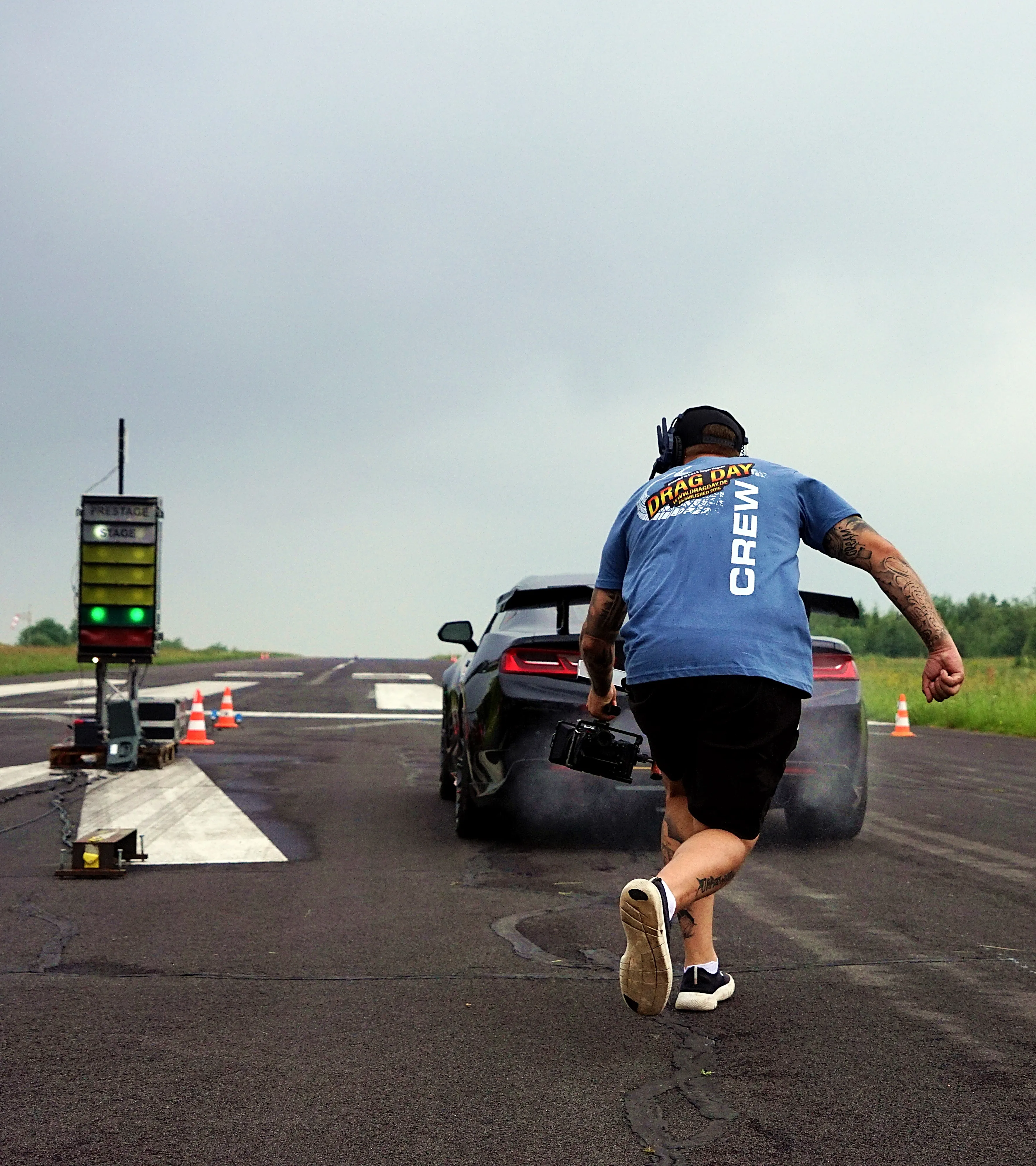 Ein Mann mit Kamera filmt ein graues Sportwagen, der auf einer Rennstrecke Rauchwolken hinterlässt. Im Hintergrund eine digitale Anzeigetafel und orangefarbene Pylonen.