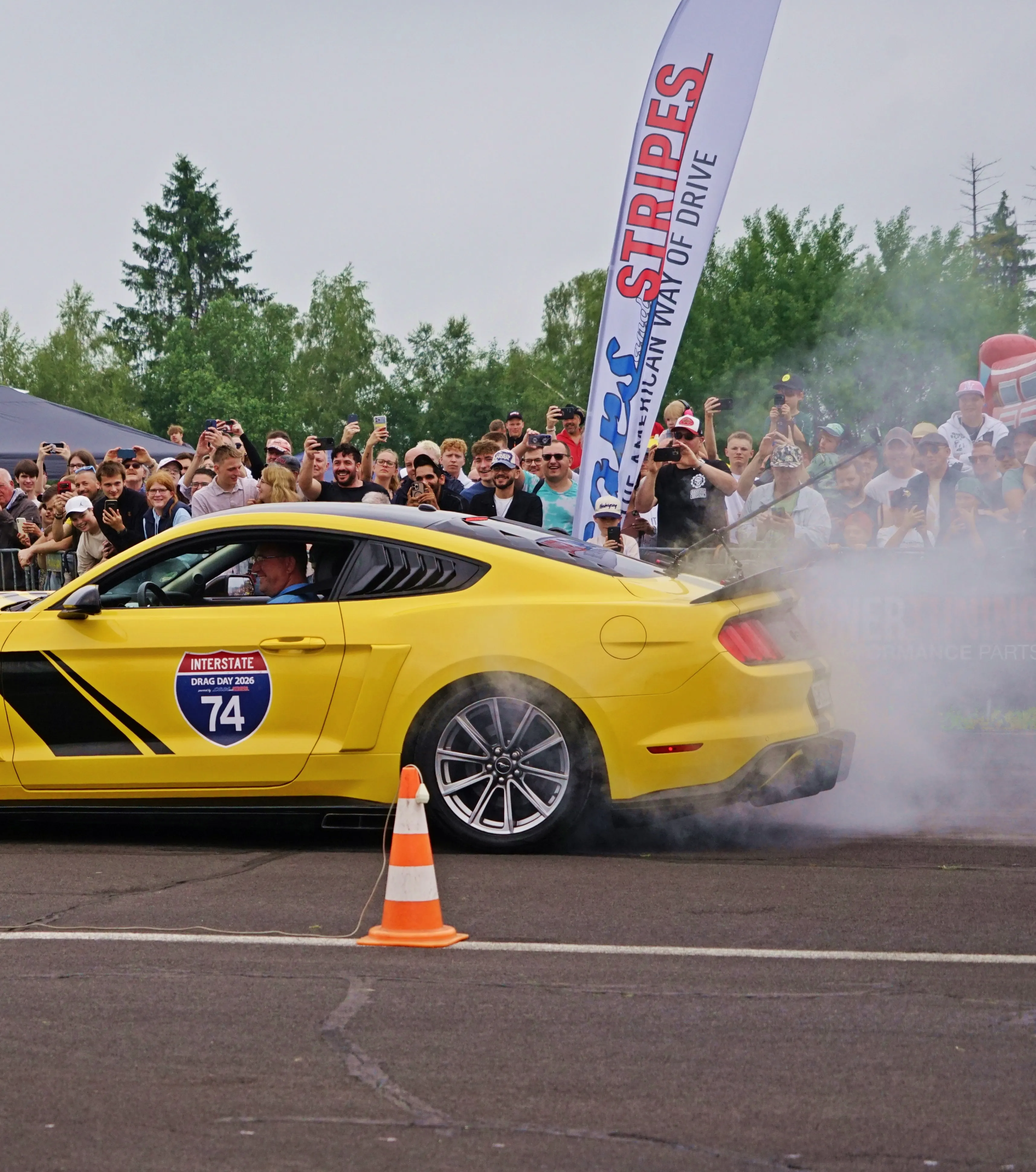 Gelber Sportwagen mit dem Aufkleber „Interstate 74 Drag Day 2026“ erzeugt Rauch beim Beschleunigen vor einer jubelnden Menschenmenge.