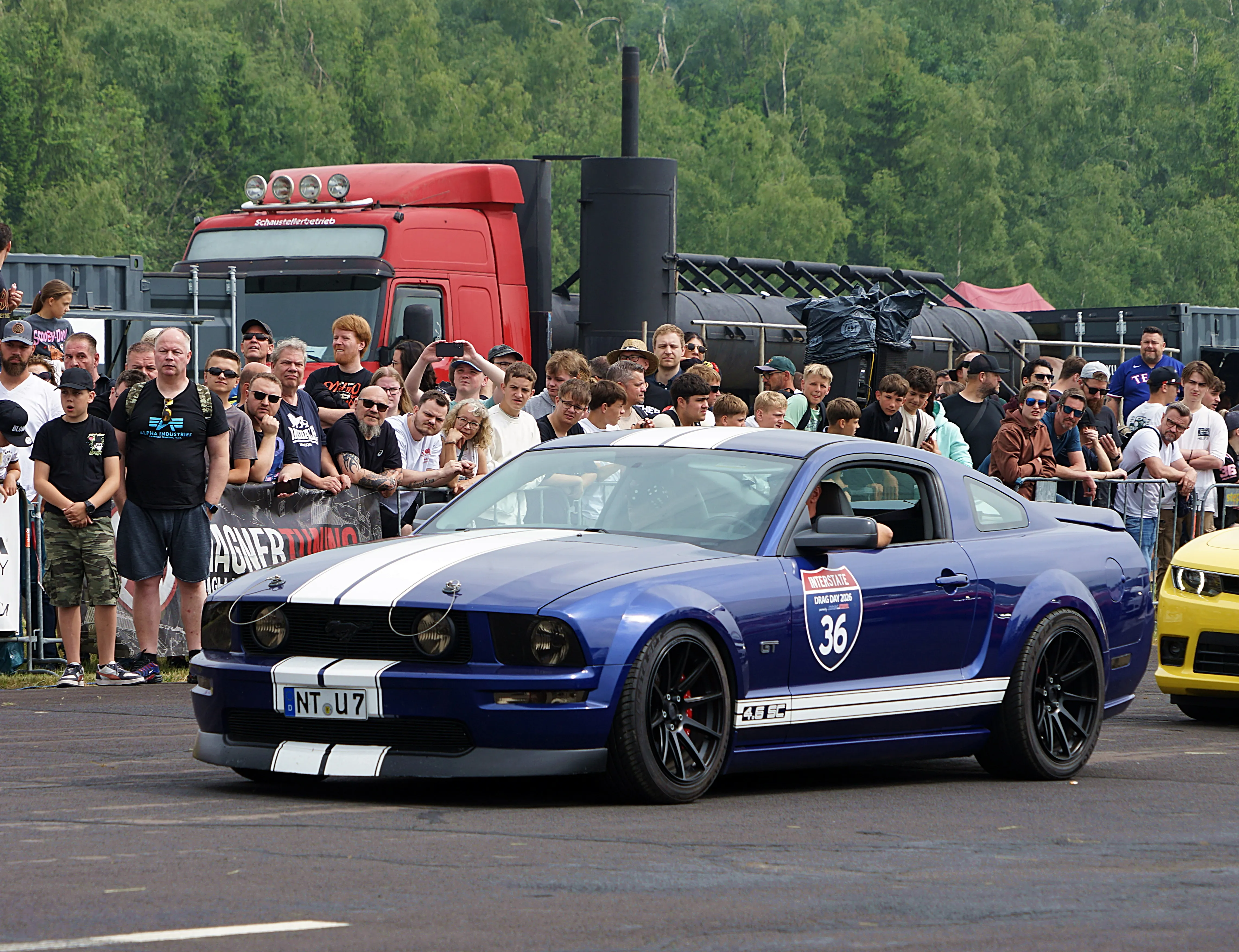 Blauer Ford Mustang mit weißen Streifen steht auf einer Straße, umgeben von Zuschauern und einem roten LKW im Hintergrund.