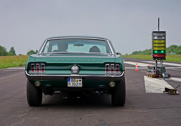 Grüner Ford Mustang steht auf einer Rennstrecke, bereit für den Start. Im Hintergrund eine Startampel und ein Feld.