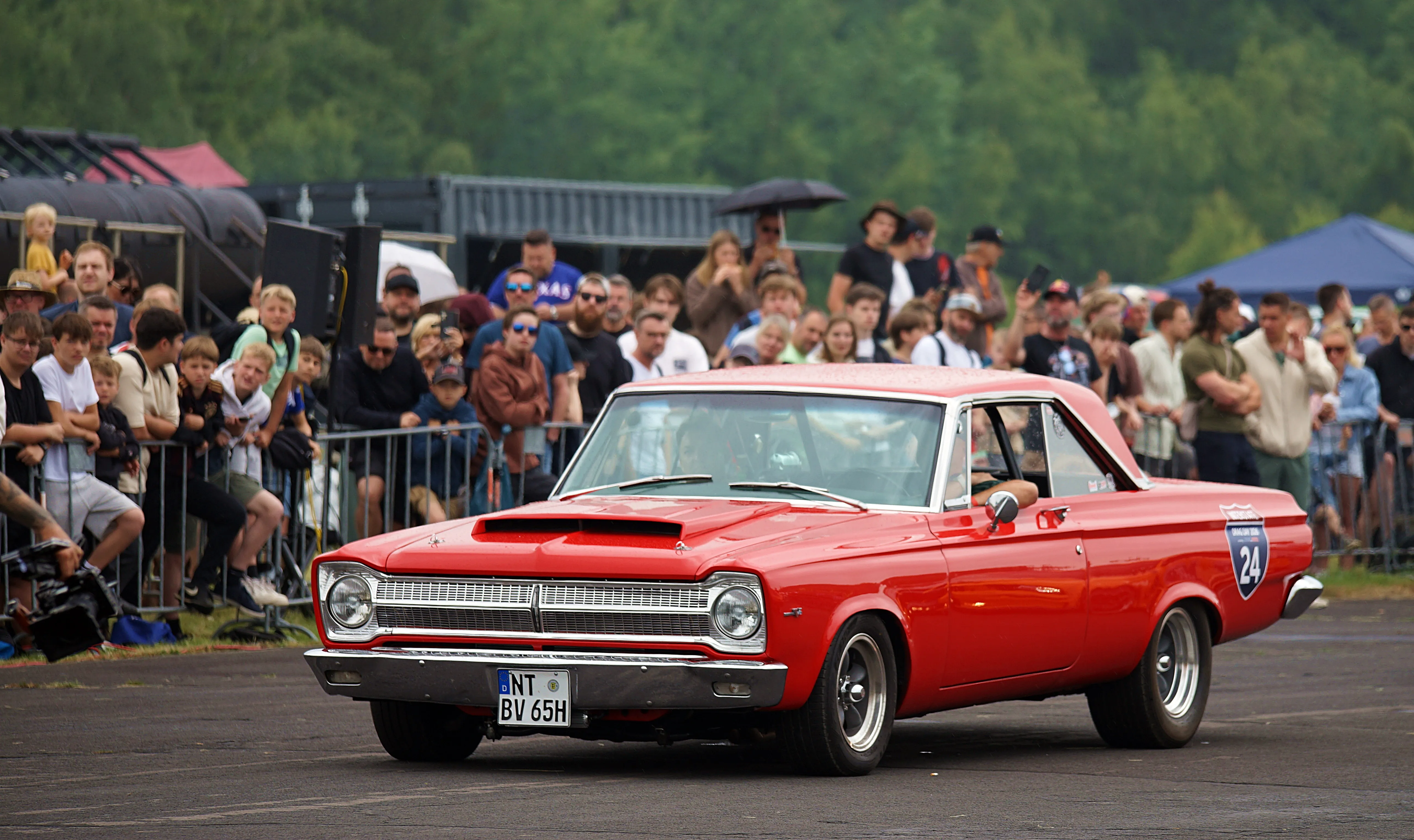Roter Oldtimer steht auf einer Rennstrecke, umgeben von Zuschauern hinter einer Absperrung.