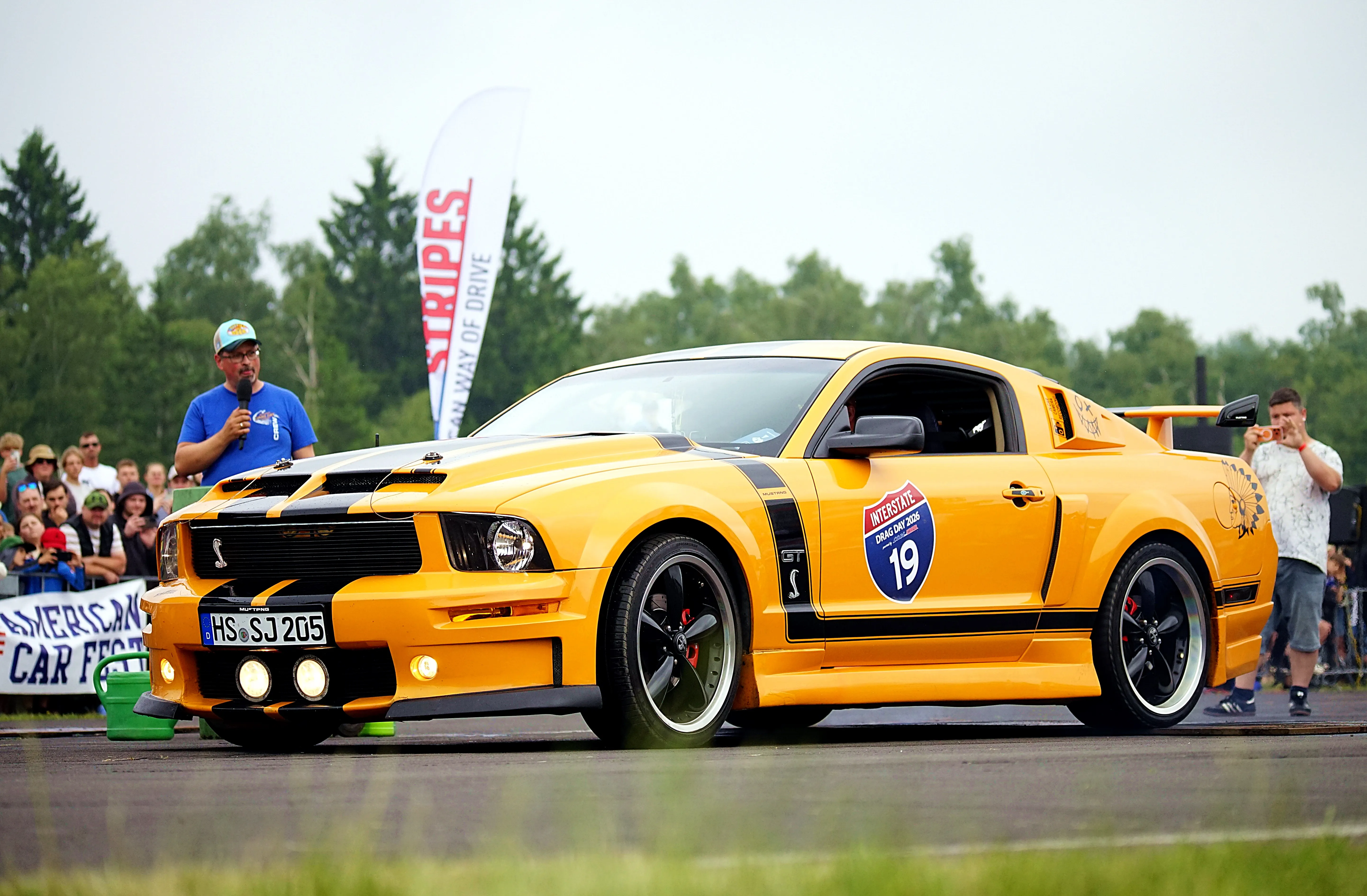 Gelber Ford Mustang mit Startnummer 19 steht auf einer Rennstrecke, umgeben von Zuschauern und Werbebannern.
