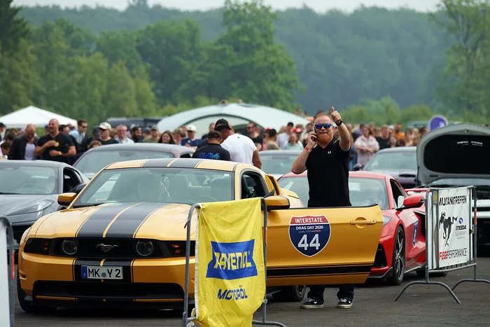 Ein Mann steht auf dem Dach eines gelb-schwarzen Ford Mustang und zeigt den Daumen nach oben. Im Hintergrund sind weitere Autos und eine Menschenmenge zu sehen.